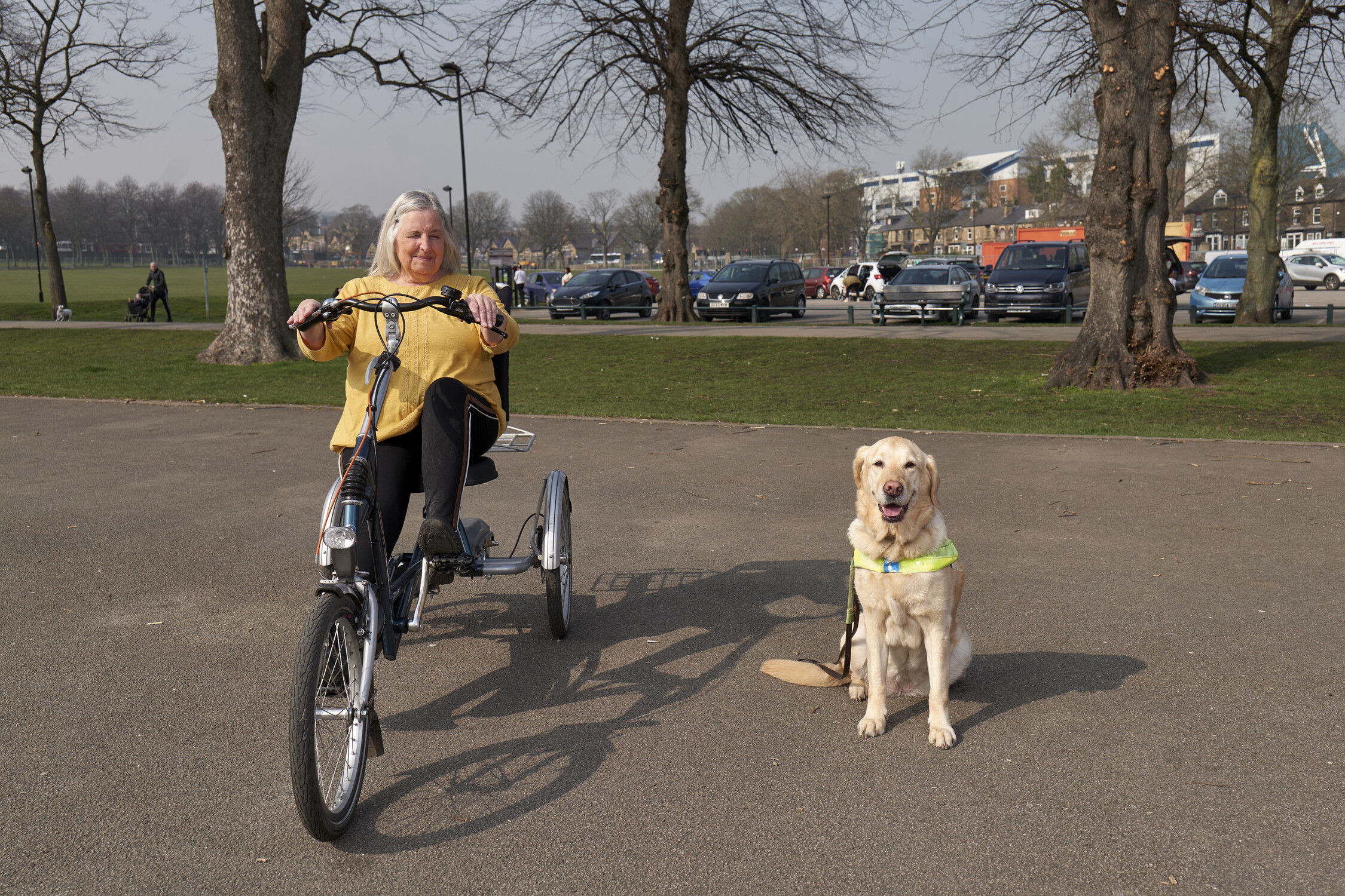 Older woman wearing yellow top and black trousers is sitting on a tricycle and there is a guide dog on the right sat on the floor