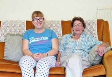 The image shows two women sat on a couch together. The woman on the right is older than the woman on the left. They are both smiling at the camera.