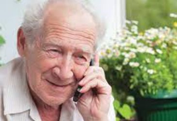 Image is of an older man with a phone held to his ear. He is smiling and there are flowers in the background.