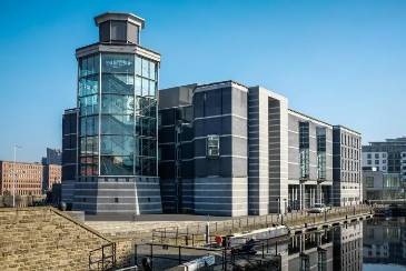 An image of the Royal Armouries Museum. In the foreground is Leeds Dock and a canal barge is visible. The background is a bright blue sky. To the left of the museum, it's winding staircase looks like a tower.