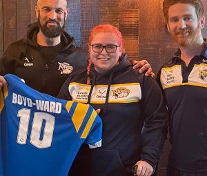 Jodie is stood wearing a Leeds Rhinos hoodie, holding a Leeds Rhinos shirt that says 'Boyd-Ward 10' on it. Jodie is stood in between two people who are also wearing Leeds Rhinos coats.