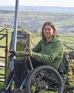 Photo shows Craig Grimes. Craig is in a wheelchair in the countryside. Craig is in front of a dry-stone wall with the word 'Heath' on it. There is also a metal gate to his left.
