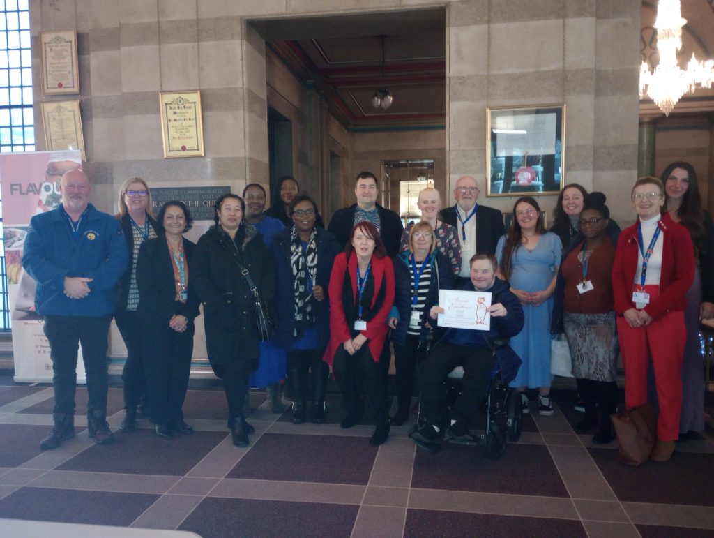Image shows the Armley Helping Hands team with their certificate. They are all smiling in front of the camera. They are stood in Leeds Civic Hall.