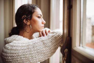 Image shows a women in a beige jumper looking out of a window. She looks worried.