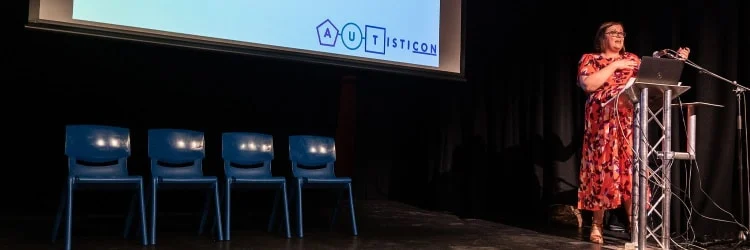 Image shows someone speaking at a lectern. A presentation is viewable behind them. It says Autisticon.